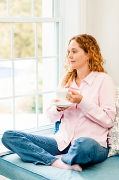 Smiling caucasian woman relaxing on couch by window holding cup of coffee