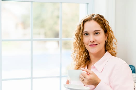 Smiling caucasian woman relaxing by window holding cup of coffee
