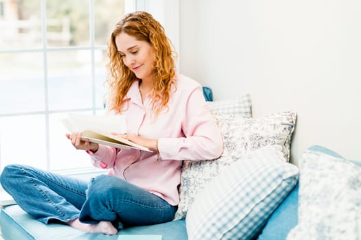 Smiling caucasian woman reading book by window at home