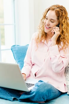Smiling caucasian woman using laptop computer while on cellphone at home