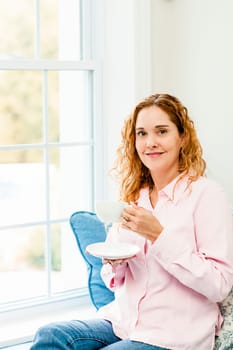 Smiling caucasian woman relaxing on couch by window holding cup of coffee