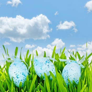 Three blue speckled easter eggs with ribbons in green grass on sky background