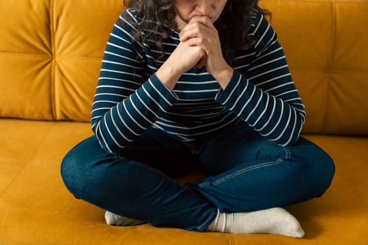 A beautiful woman with dark long hair in a striped sweater and blue jeans sits on the sofa with her legs crossed cross-legged and her chin resting on her hands. Girl resting on a yellow sofa