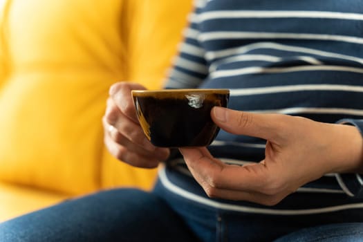 An unrecognizable woman is sitting on the sofa and holding a cup of coffee in her hands. Unrecognizable happy girl holding a mug of hot coffee. girl drinks a hot drink with pleasure