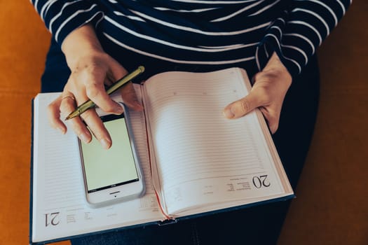 a female entrepreneur at work. Work at home: A business woman uses a smartphone and takes notes. the girl makes notes in a notebook while working with a smartphone. green blank screen of her smartphone. empty space for advertising and copy