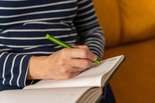 an unrecognizable woman, writing notes with a pen in a notebook, sitting on the couch. All her attention is focused on capturing every thought, inspiration or new idea.