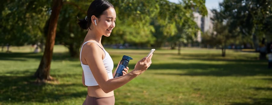 Portrait of fitness girl, runner drinking water, looking at smartphone, standing in park in middle of workout, sport training session.