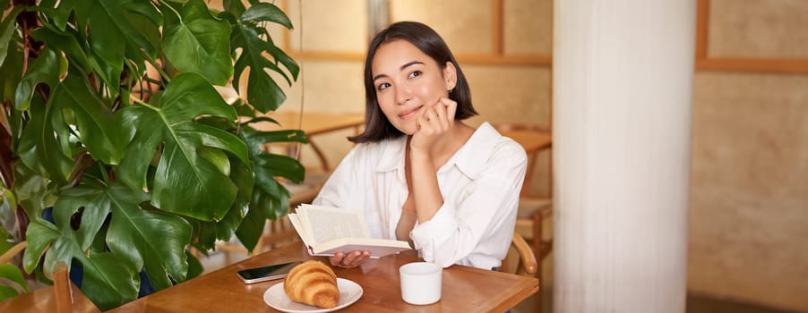 Dreamy young smiling asian woman reading book, sitting in cafe, eating croissant and drinking coffee in cozy interior.