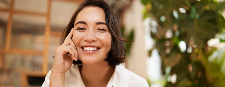 Close up portrait of funny young asian woman, sitting in cafe, chatting lively, smiling at camera.