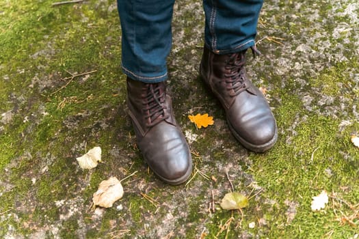 women's feet in leather autumn boots and jeans stand on a large boulder covered with green forest moss. Walking in the woods. Nature and active recreation