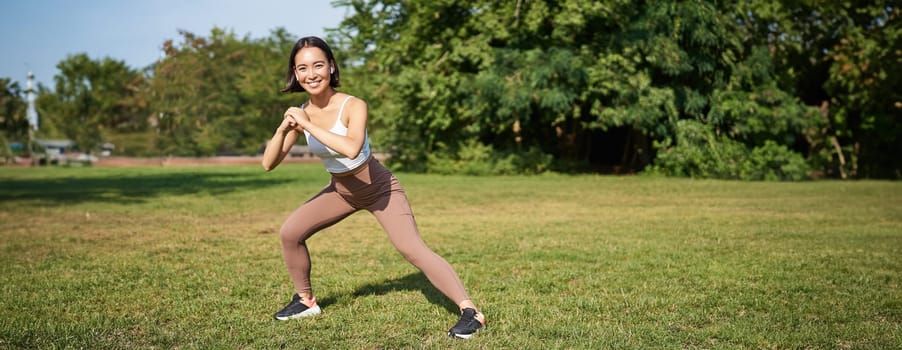 Wellbeing and sport complex. Young asian woman stretching, doing squats and workout on fresh air, smiling pleased.