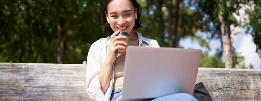 Asian girl sitting with laptop and wireless earphones, drinking coffee, looking at screen, doing homework, working on remote.