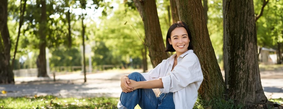 Stylish young university student, girl sits in park on lawn, leans on tree and smiles, resting outdoors and enjoying nature.