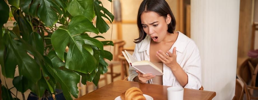 Portrait of asian woman looking shocked at book pages, reading something interesting, concentrating, sitting in cafe with coffee and croissant.