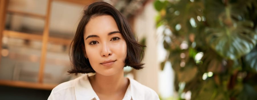 Close up portrait of beautiful, romantic young asian woman, smiling and looking happy, sitting in cozy cafe. Copy space