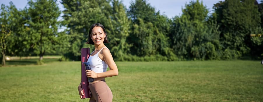 Vertical shot of fit and healthy asian woman posing in park, holding water bottle and yoga rubber mat for workout outdoors.