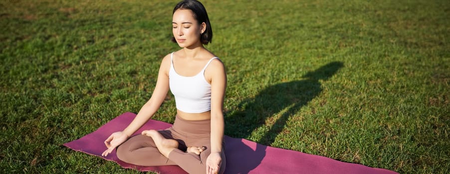 Mindfulness and meditation. Young asian woman smiling while doing yoga, relaxing in asana on rubber mat, doing exercises in park on fresh air.
