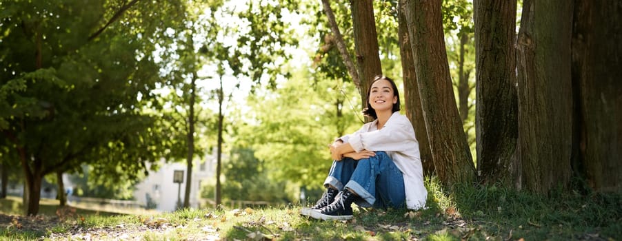 Portrait of beautiful asian woman resting near tree, relaxing in park, smiling and looking happy. Copy space