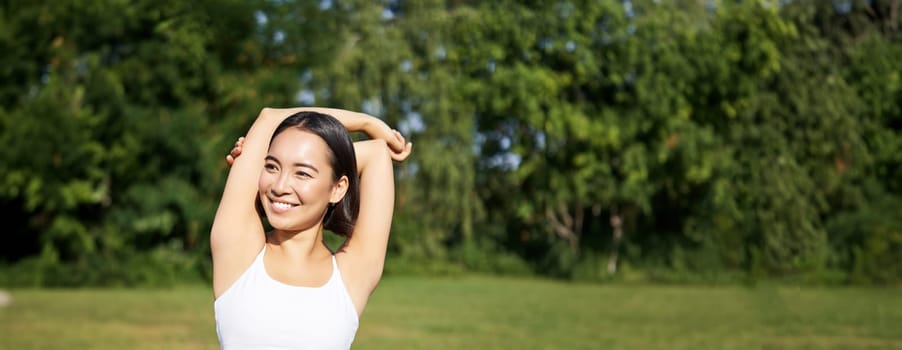 Asian woman stretching her arms, doing fitness workout in park, smiling pleased, warming up before jogging on fresh air in daytime.