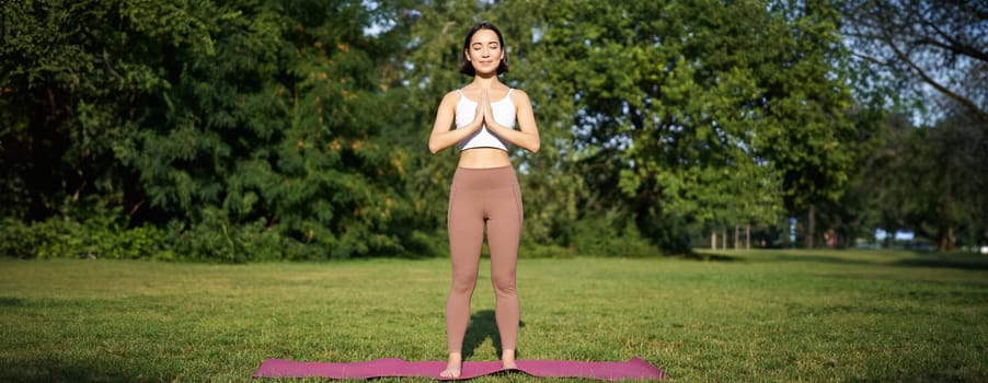 Young asian woman in sportswear, holding hands in namaste gesture, practice yoga, meditating and relaxing on fresh air in park, concept of mindfulness and wellbeing.