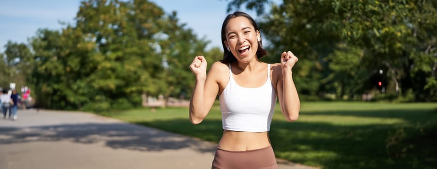 Happy fitness girl achieve goal, finish marathon, running with hands up, celebrating victory while jogging, triumphing in park.