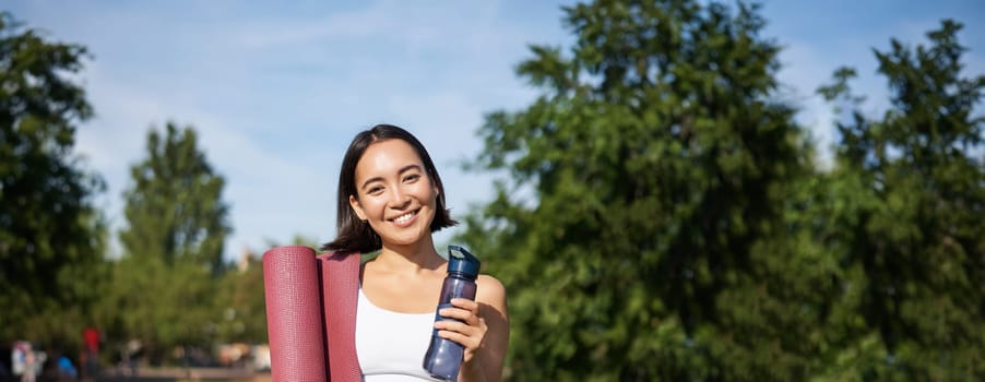 Portrait of young slim and healthy korean girl doing workout in park, standing with water bottle and rubber mat for execises on green lawn, smiling happily.