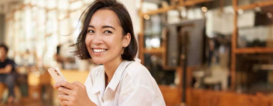 Vertical portrait of stylish asian woman sitting in cafe, drinking coffee and using smartphone. Lifestyle and people concept