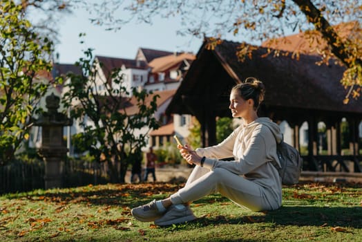 Young fashionable teenage girl with smartphone in park in autumn sitting at smiling. Trendy young woman in fall in park texting. Retouched, vibrant colors. Beautiful blonde teenage girl wearing casual modern autumn outfit sitting in park in autumn. Retouched, vibrant colors, brownish tones.