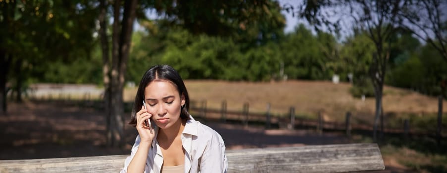 Portrait of sad girl talking on mobile phone and sulking. Young woman having an uneasy conversation on telephone, sitting on bench in park with upset face.