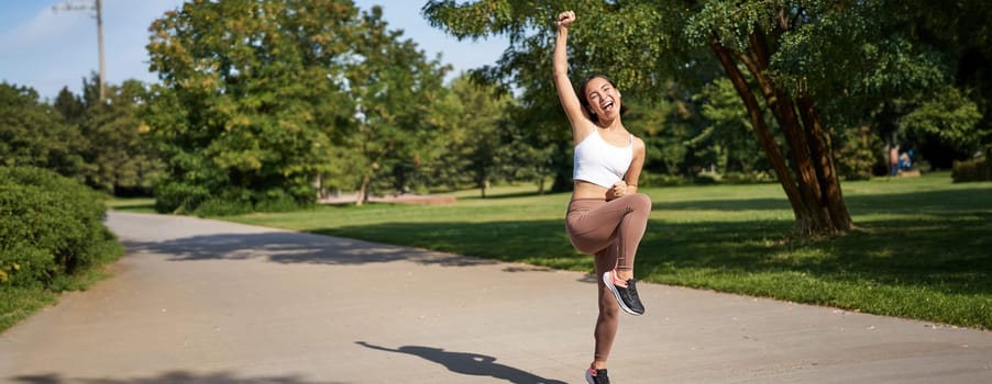 Excited young asian woman winning, finish running in park, saying yes, lifting hand up in triumph, celebrating victory or success.