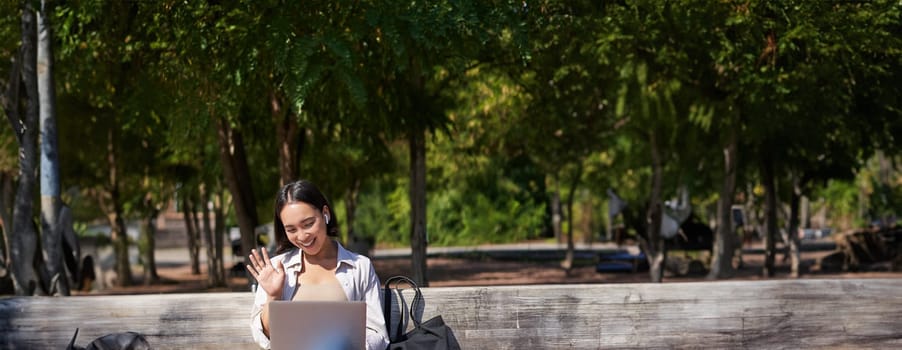 Portrait of asian woman sitting with laptop on bench in park, listening music with wireless headphones, doing homework, working on remote.