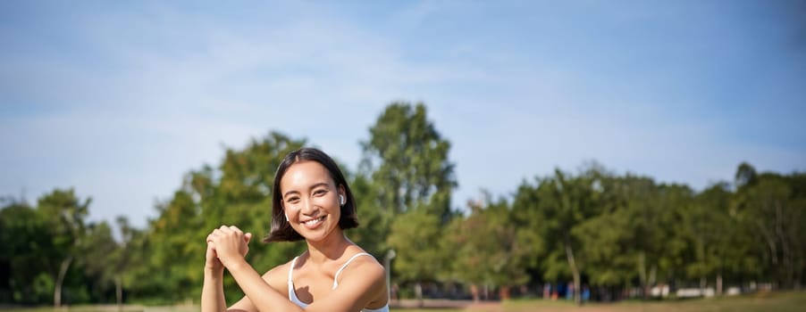 Vertical shot of young fit woman does squats in park, using stretching band on legs, smiling pleased while workout.