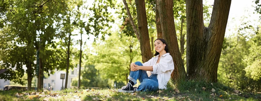 Beautiful smiling girl sits near tree in park, enjoying nature outdoors, relaxing and resting on fresh air. Copy space