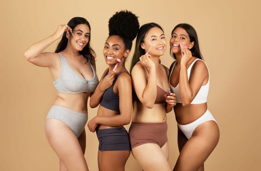 Young diverse women in underwear confidently apply serum and use beauty tools, showcasing their natural beauty against beige background, smiling at camera