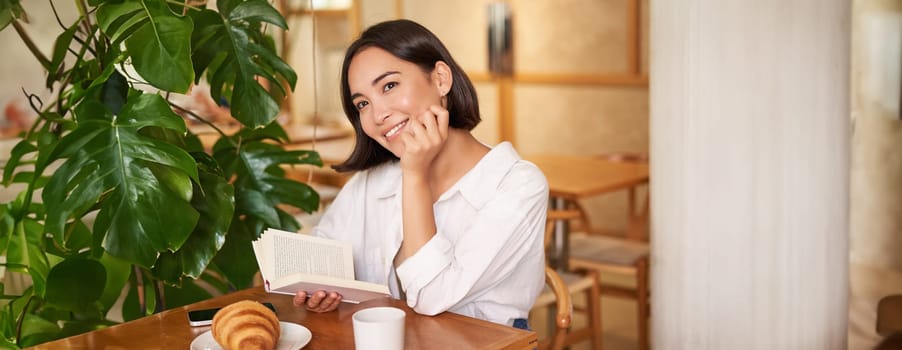 Romantic asian woman sitting with book in cafe, eating croissant and drinking coffee, reading and smiling, enjoying alone time.