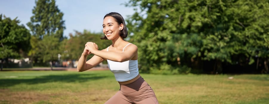 Wellbeing and sport complex. Young asian woman stretching, doing squats and workout on fresh air, smiling pleased.