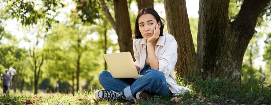 Portrait of young asian woman sitting in park near tree, working on laptop, using computer outdoors.