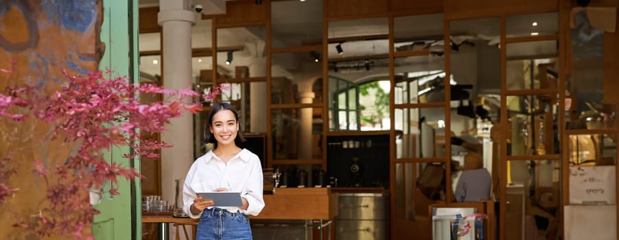 Portrait of asian woman, manager standing with tablet in front of cafe entrance, welcomes guests. Business and entrepreneurs concept