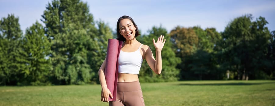 Portrait of young asian woman workout in park outdoors, fitness instructor waves hand and smiles, invites people for yoga training on fresh air.