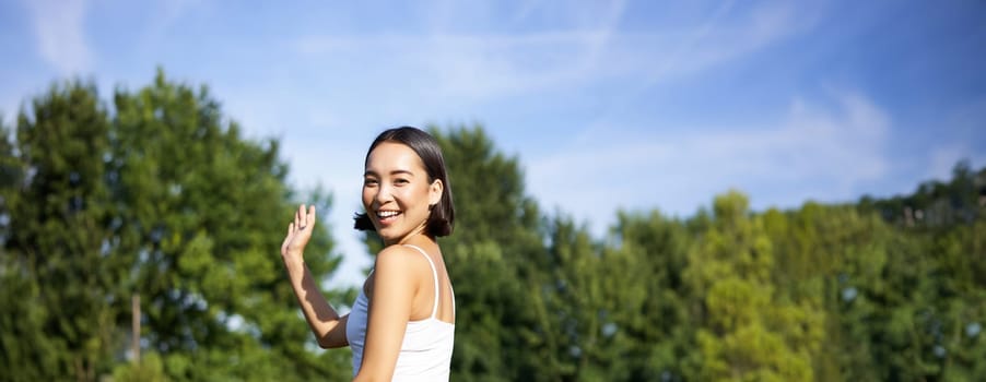 Portrait of asian girl on fitness class in park, sitting on rubber mat and wave hand at camera, say hello, meditating and practice yoga.