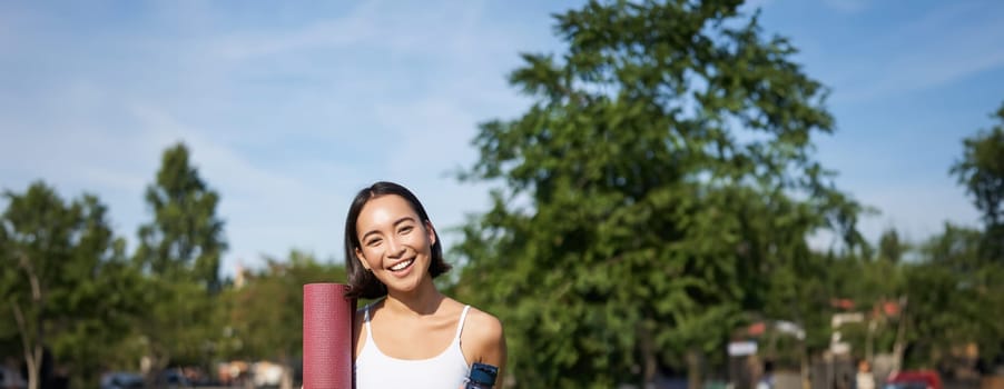 Portrait of young slim and healthy korean girl doing workout in park, standing with water bottle and rubber mat for execises on green lawn, smiling happily.