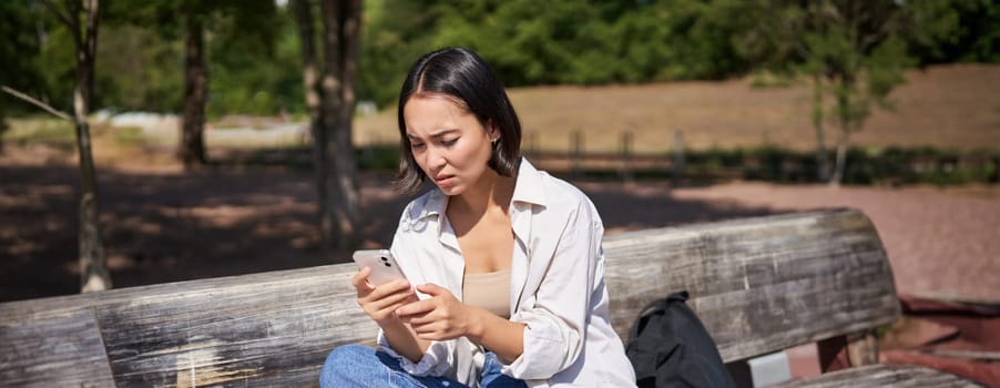 Asian girl frowning, looking concerned at her mobile phone, reading sad message, bad news on smartphone app, sitting on bench in park worrying.