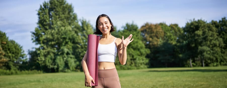 Portrait of young asian woman workout in park outdoors, fitness instructor waves hand and smiles, invites people for yoga training on fresh air.