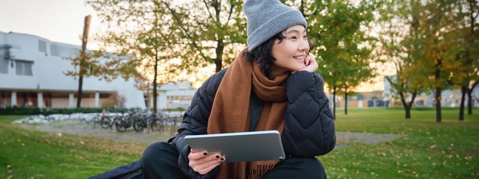 Smiling asian girl, student sits on bench in park alone, reading, using social media app on digital tablet, watches videos outdoors, relaxes on fresh air.