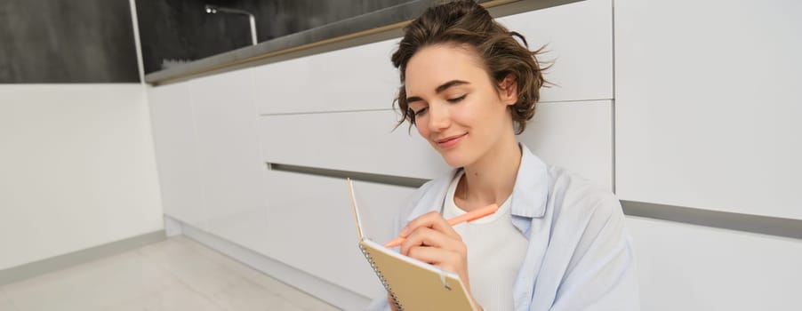 Portrait of beautiful young woman writing in journal, adding notes in planner, sitting on floor and thinking, reads her diary, smiles with pleased face expression.