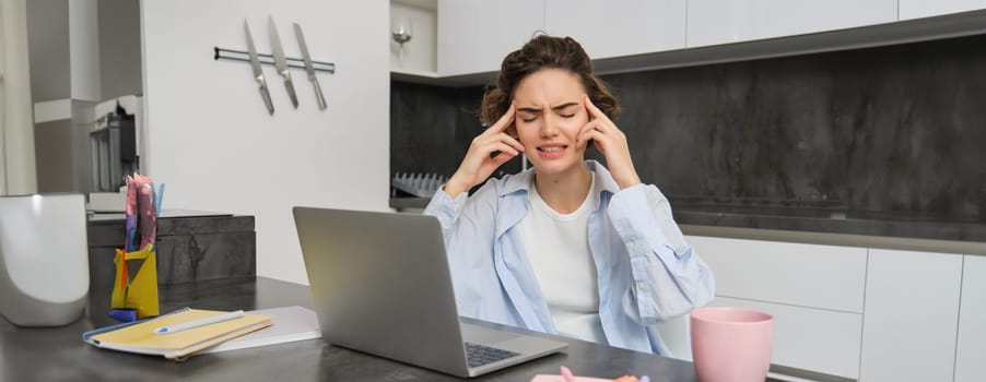 Portrait of troubled young woman, sits near laptop with frustrated face expression. Girl suffers migraine while works on remote, touches head and grimaces from headache.