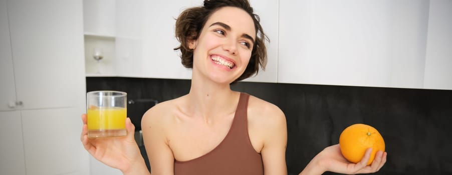 Healthy lifestyle and sport. Beautiful smiling woman, drinking fresh orange juice and holding fruit in her hand, enjoying her vitamin drink after workout at home, standing in kitchen.