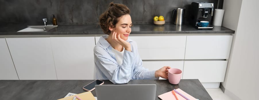 Beautiful woman managing her work from home, drinking coffee and looking at laptop, sitting in kitchen with paperwork, doing her taxes, studying indoors on remote.