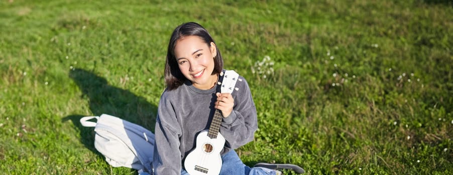 Smiling asian girl learns how to play ukulele on laptop, video chat with music teacher, sitting with instrument in park on grass.