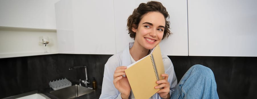 Portrait of smiling young woman reading journal, enjoys comfort at home, holding notebook, looking happy, relaxing indoors.